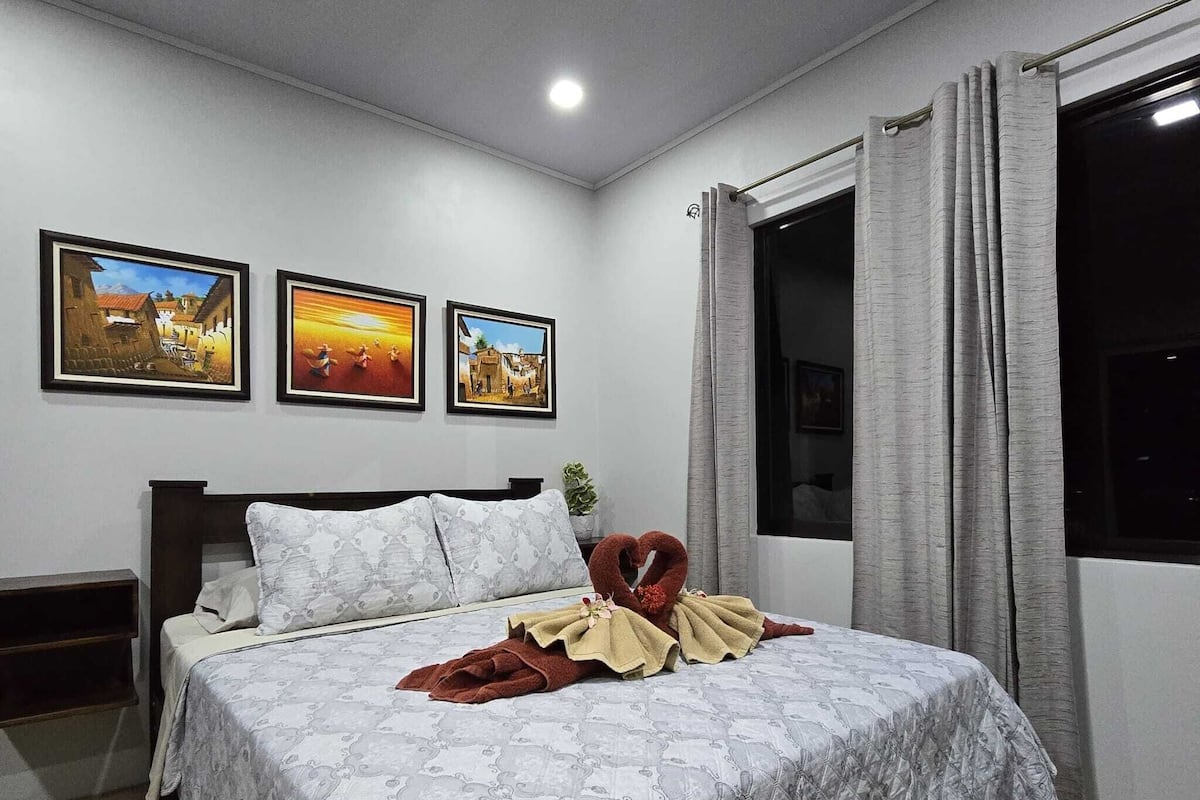 A bedroom with a bed, a nightstand, a window with curtains, and three framed pictures on the wall.