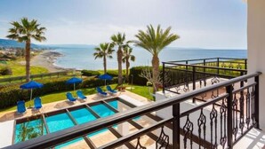 A balcony with a pool, blue umbrellas, and a view of the sea and beach. - Villa Pelagos (Nea Dimmata)
