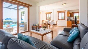A spacious living area with a sofa, a coffee table, and a dining table with chairs. There is a sliding glass door leading to a balcony with a pool view. - Villa Pelagos (Nea Dimmata)