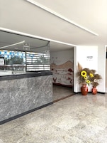 Uma área de recepção com balcão de mármore, um mural na parede e dois girassóis em vasos.