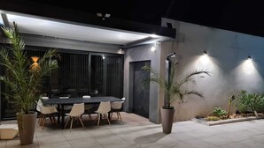 Un área de comedor al aire libre con una mesa y sillas, plantas en macetas y una fachada de edificio moderna.