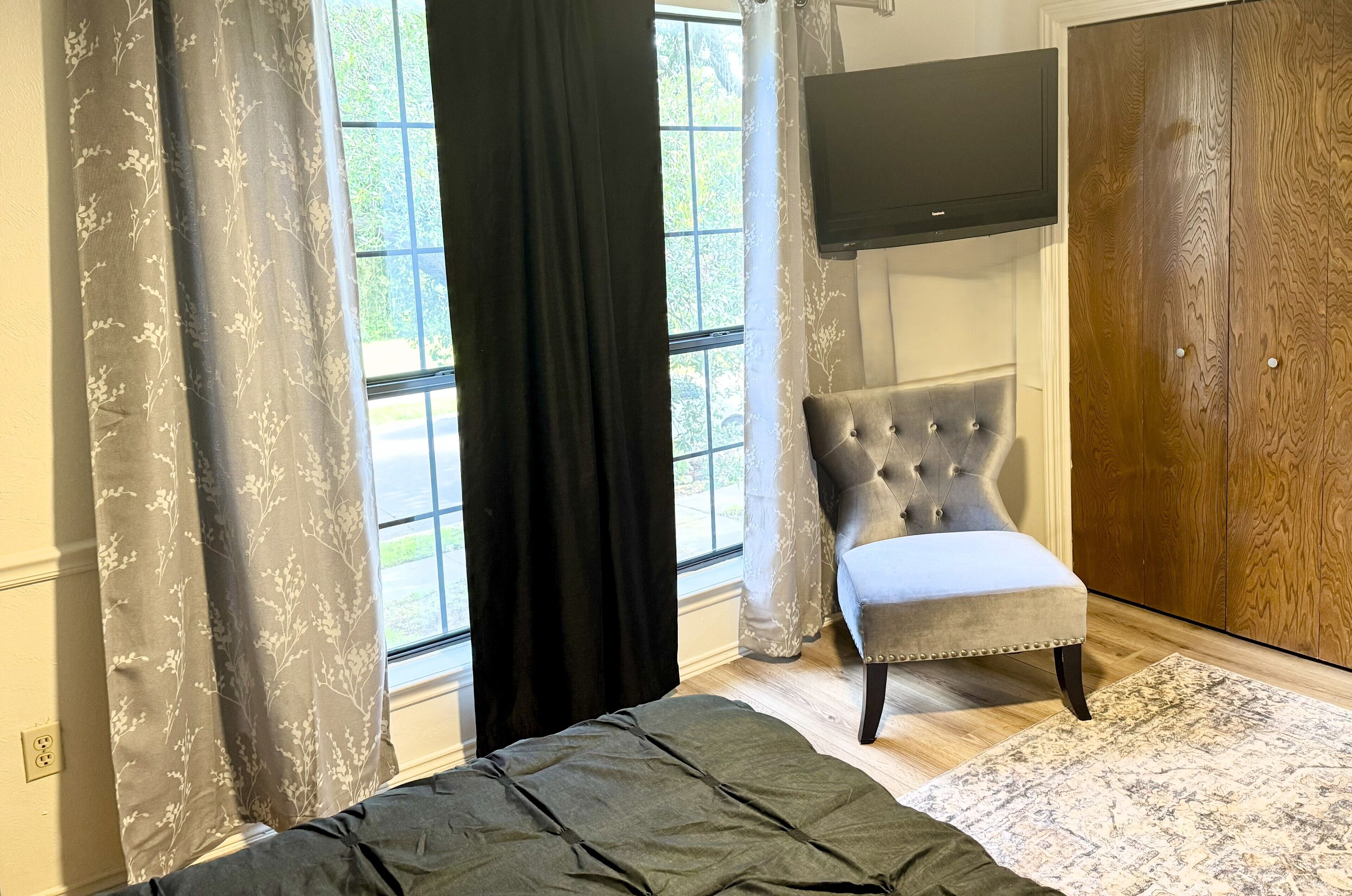 Een slaapkamer met een bed, een grijs gestoffeerde fauteuil, een houten garderobe en een aan de muur bevestigde televisie.