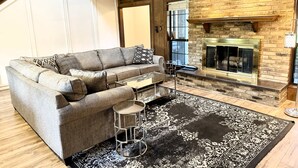 A modern living room with a grey sectional sofa, a glass coffee table, and a patterned rug. A fireplace with a brick wall and a wooden ceiling is visible in the background. - Cozy Pool Home Designed for Both Family Fun & Serenity (Plano)