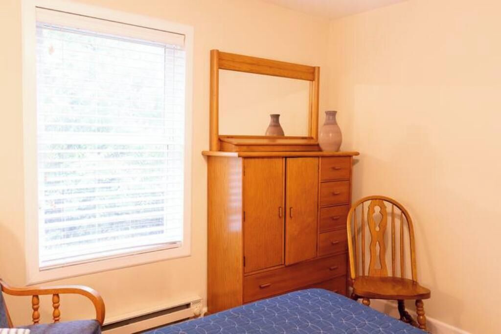 A bedroom with a wooden wardrobe, a bed with a blue bedspread, a wooden chair, and a window with blinds.