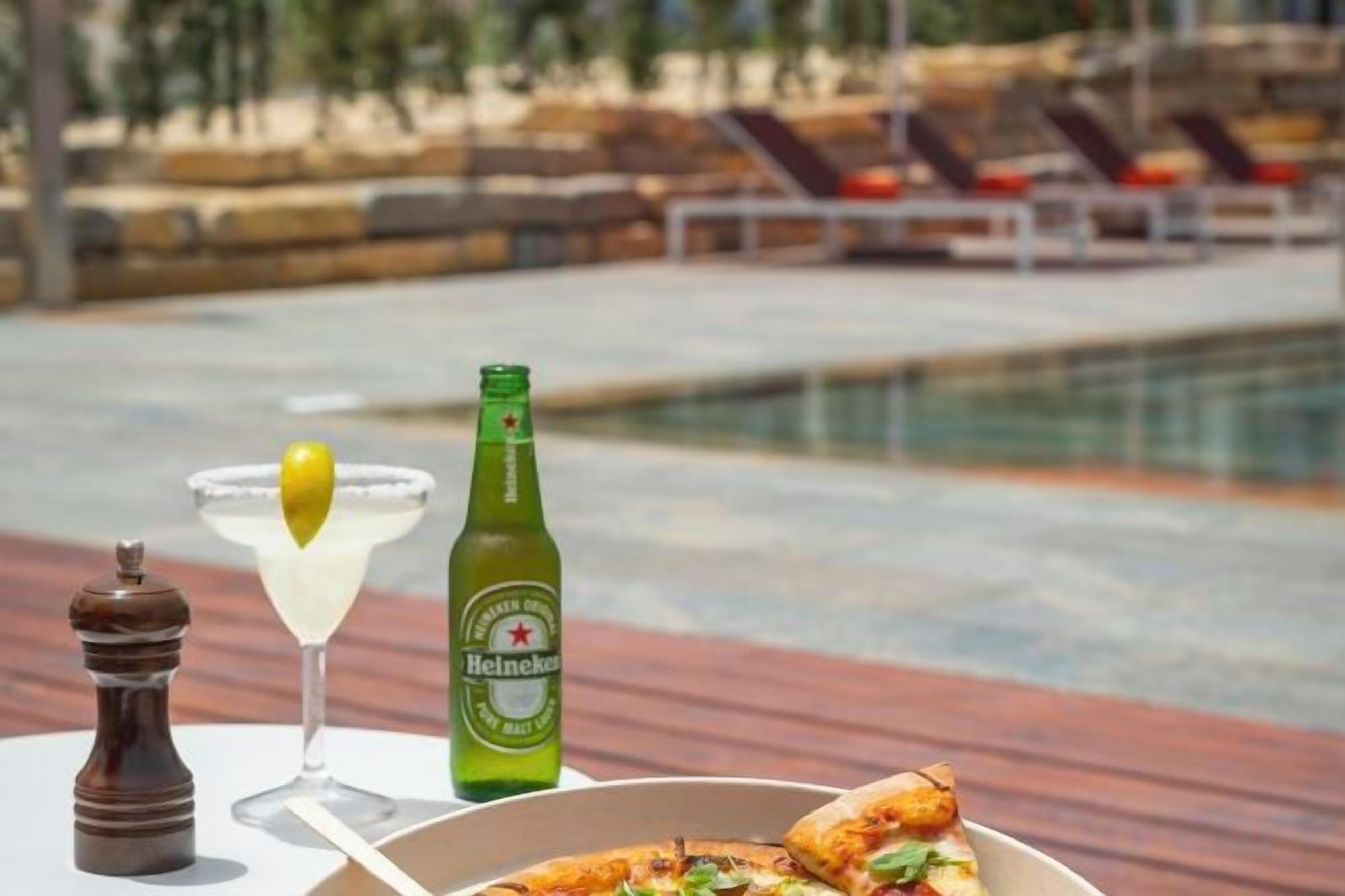 Ein Tisch mit einer Bierflasche, einem Cocktailglas und einer Pizza mit Blick auf einen Pool.