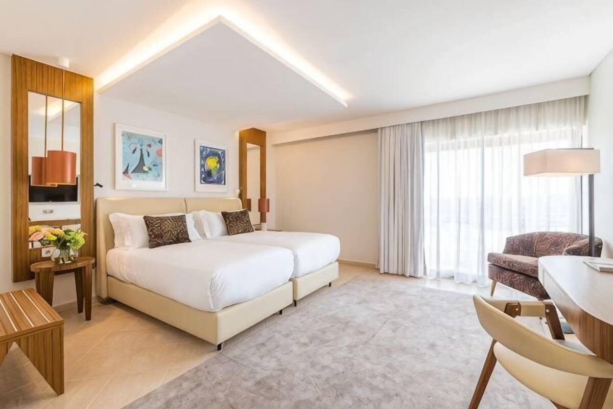A hotel room with a double bed, a desk, a chair, a small table with a lamp, and a large window with curtains.