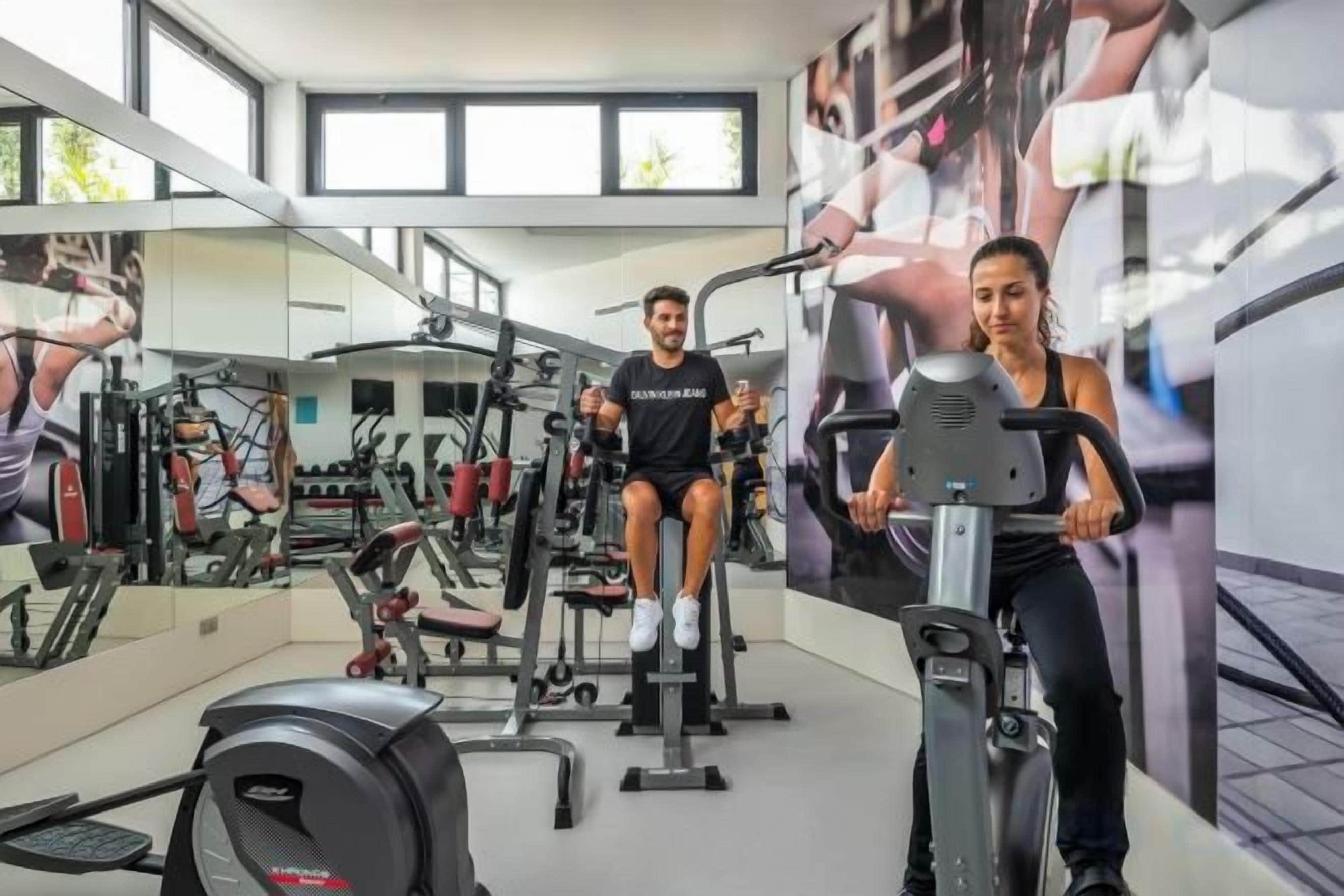 Un gimnasio con equipo de ejercicio, espejos y amplias ventanas.