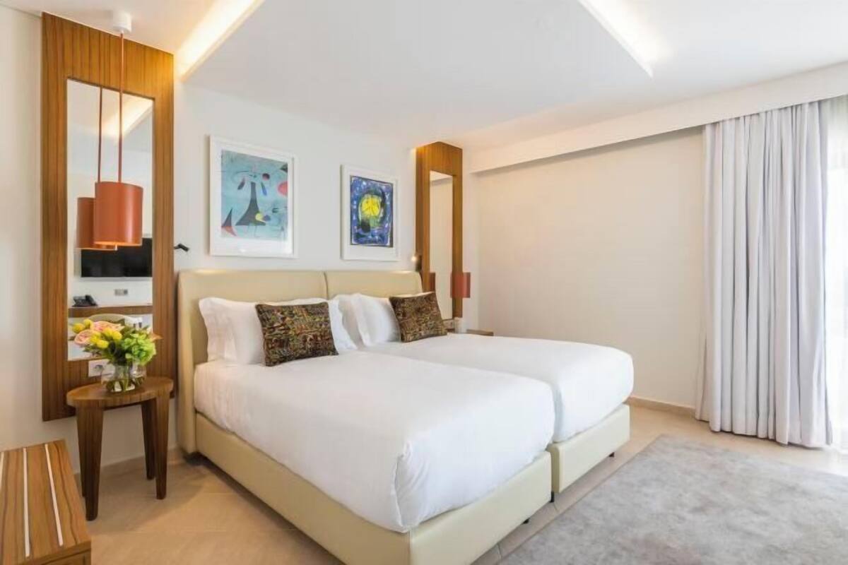 A hotel room with a double bed, a bedside table with a vase of flowers, a mirror, and two framed artworks on the wall.