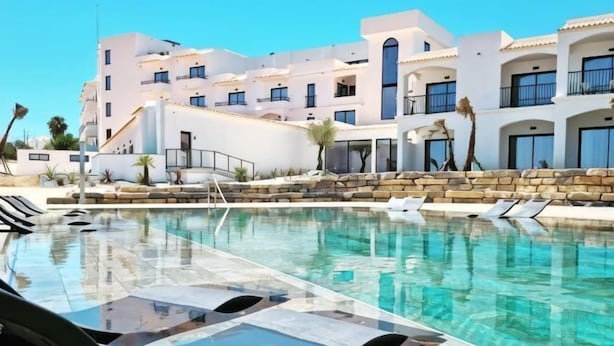 A swimming pool with lounge chairs, a clear blue sky, and a building with white walls and multiple balconies.