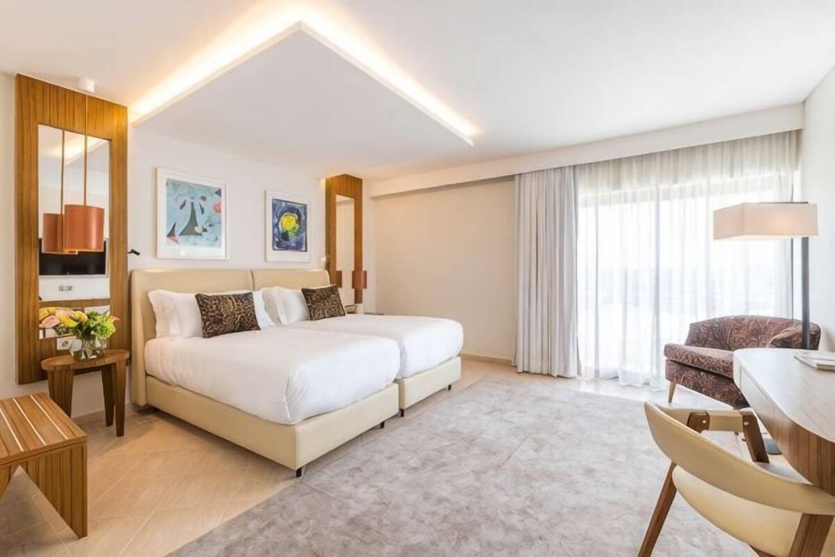 A hotel room with a large bed, a desk, a chair, a small table, and a large window.