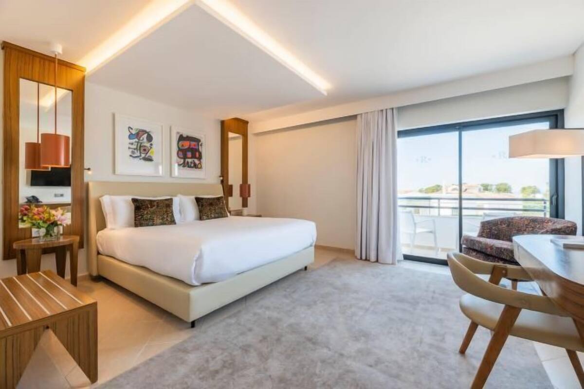 A modern hotel room with a large bed, a dining area, a balcony with a view, and artwork on the walls.