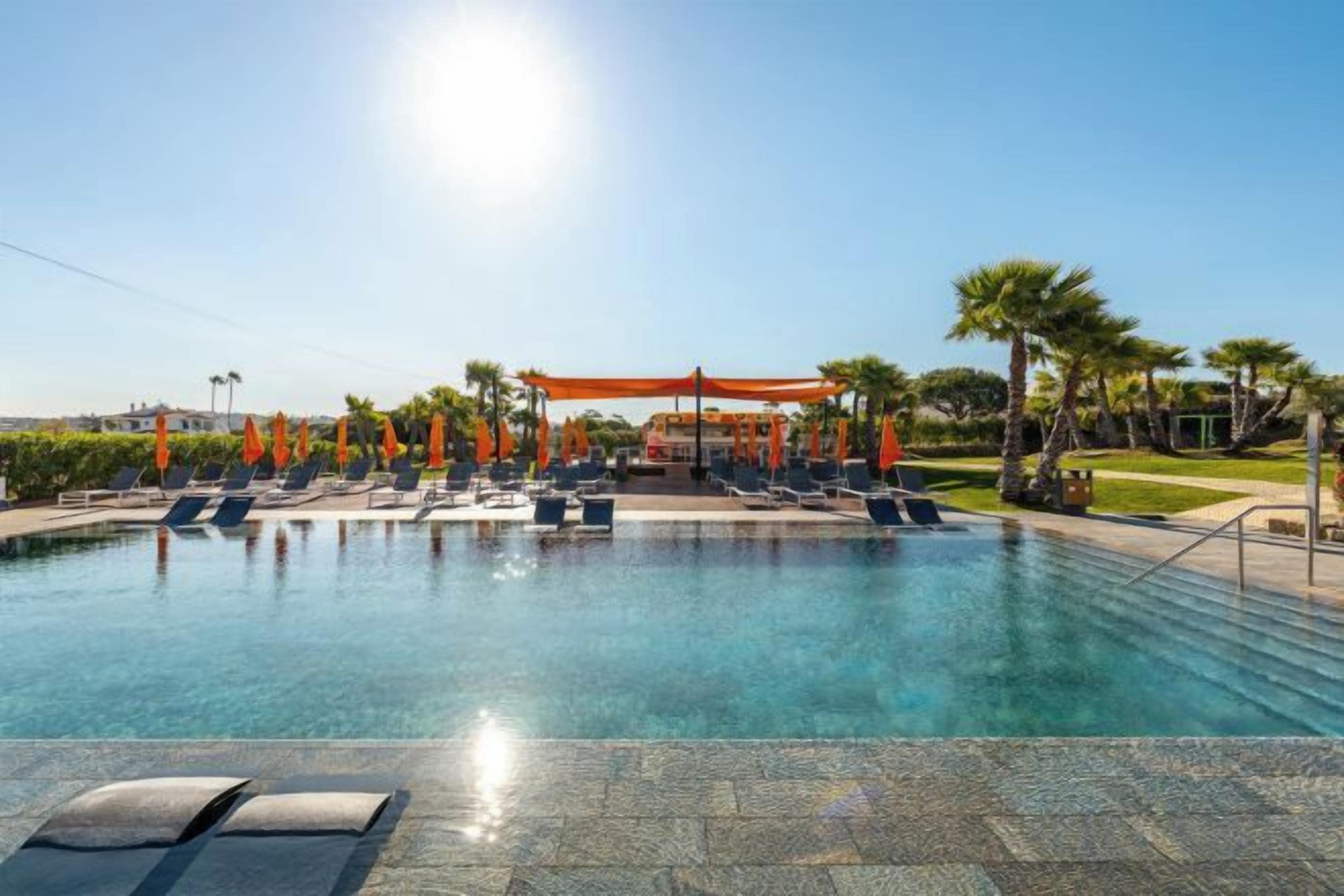 A swimming pool with lounge chairs and umbrellas, surrounded by palm trees and a clear sky.