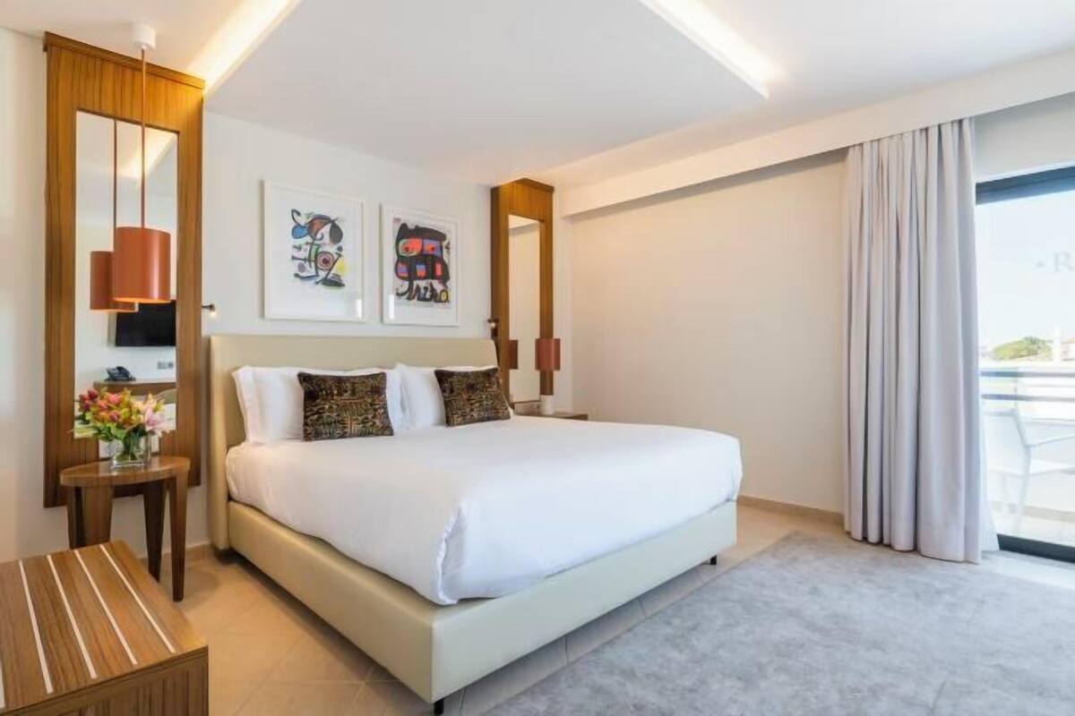 A hotel room with a large bed, a bedside table with a vase of flowers, a mirror, and artwork on the wall.