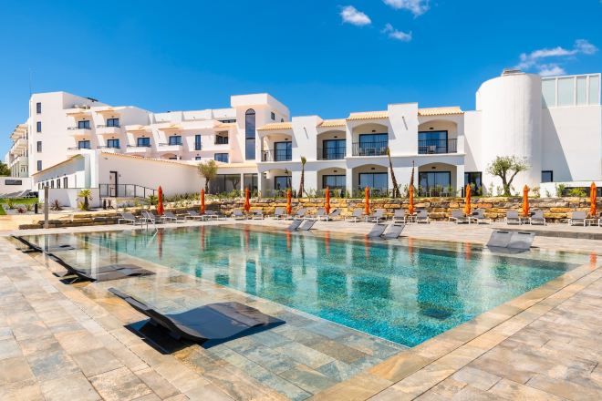A swimming pool with lounge chairs and umbrellas, a modern building with multiple windows, and a clear blue sky.
