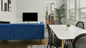 A modern living room with a flat-screen TV, blue sofa, dining table, chairs, ceiling fan, and a view of the kitchen.