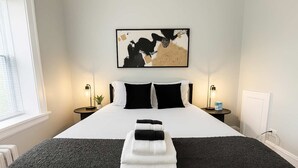 Una habitación de hotel con una cama grande, ropa de cama blanco y negro, dos mesitas de noche con lámparas, una obra de arte en la pared y una ventana con persianas.