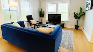 Sala de estar con sofá azul, un sillón negro, una mesa de centro, televisor sobre un mueble bajo y dos plantas en macetas.
