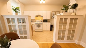 Cocina compacta con muebles blancos, un microondas y una estufa. Hay una mesa de comedor con sillas y una planta en maceta.