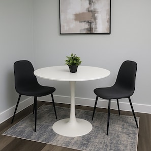 A round white table with two black chairs, a small potted plant, and a gray rug on a wooden floor.