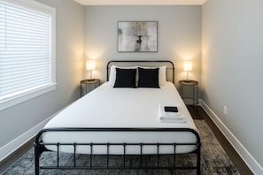 Een netjes opgemaakt bed met een witte deken en zwarte kussens, twee bedlampen, een lijst met abstracte kunst boven het bed, en een raam met jaloezieën links.