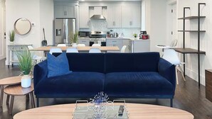 A modern living room with a dark blue sofa, a round coffee table, and a dining area with a kitchen in the background. - 6-bed, Close to City, Roof deck! Private Bathroom per Bedroom! (Boston)