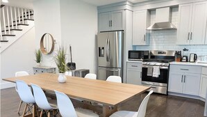 A modern kitchen with a wooden dining table, white chairs, stainless steel appliances, and a staircase in the background. - 6-bed, Close to City, Roof deck! Private Bathroom per Bedroom! (Boston)