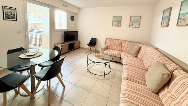 Una sala de estar moderna con un sofá a rayas, una mesa de comedor de cristal con sillas, un televisor sobre un mueble de madera y un balcón visible a través de una puerta abierta.