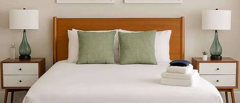 A neatly made bed with a wooden frame, green pillows, and a white duvet. There are two bedside tables with lamps and a framed picture above the bed.
