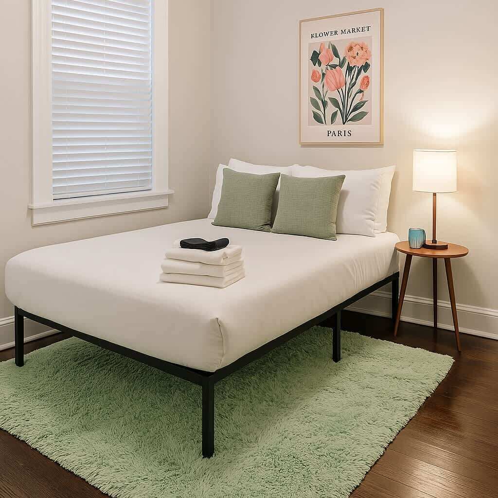 A neatly made bed with a green rug, a bedside table with a lamp, and a framed picture on the wall.