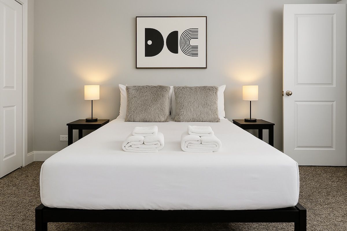 A neatly made bed with white linens and two pillows, flanked by two bedside tables with lamps, in a room with a plain wall and a door.