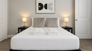 A neatly made bed with white linens and two pillows, flanked by two bedside tables with lamps, in a room with a plain wall and a door.
