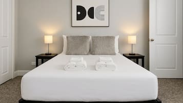 A neatly made bed with white linens and two pillows, flanked by two bedside tables with lamps, in a room with a plain wall and a door.