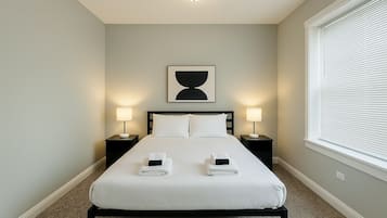 A neatly made bed with two pillows and two folded towels, a bedside table with a lamp, a framed artwork above the bed, and a window with blinds on the right.