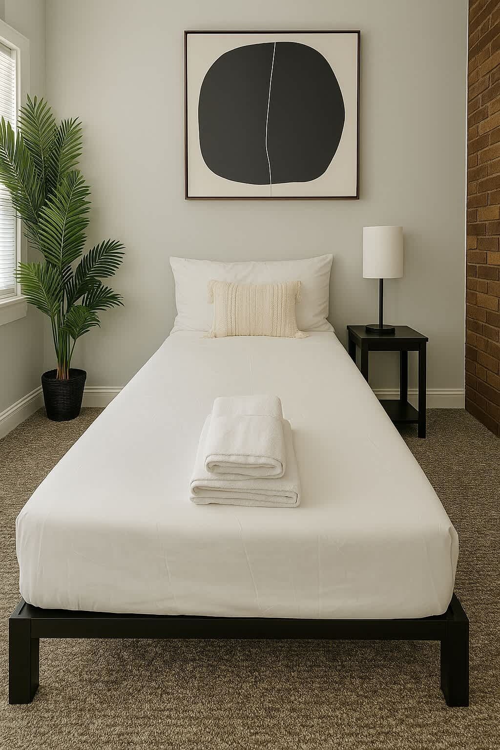 A neatly made bed with a folded white towel, a potted plant, a wall-mounted abstract painting, and a nightstand with a lamp.