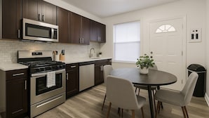A modern kitchen with dark cabinets, stainless steel appliances, a round dining table with chairs, and a window with blinds.
