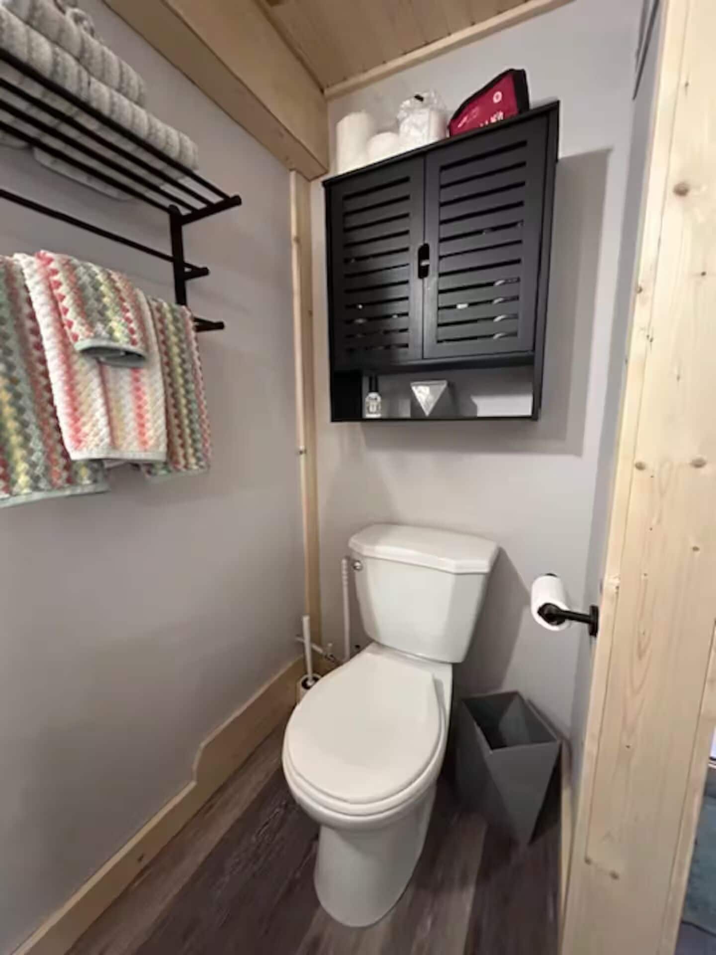 Un baño pequeño con inodoro, un cubo de basura y un perchero con toallas de colores.