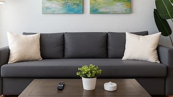 Een moderne woonkamer met een grijze bank, twee witte kussens, een kleine tafel met een plant, een afstandsbediening en een bijzettafeltje.