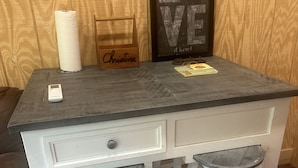 Un petit bureau en bois blanc, accompagné de deux tabourets, d’un tableau encadré, d’un porte-rouleau d’essuie-tout et d’un petit appareil électronique.