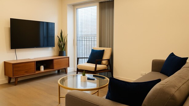 Ein modernes Wohnzimmer mit Flachbildfernseher, einer Holzkonsole, einem runden Glastisch und einem beige farbenen Sofa mit blauen Kissen.