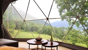 A cozy room with a large window, a bowl of fruit, a bottle of wine, and two glasses on a small table.