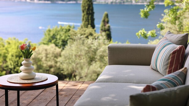 A sofa with striped pillows and a vase with flowers on a wooden table, overlooking a lake with trees and buildings in the background.