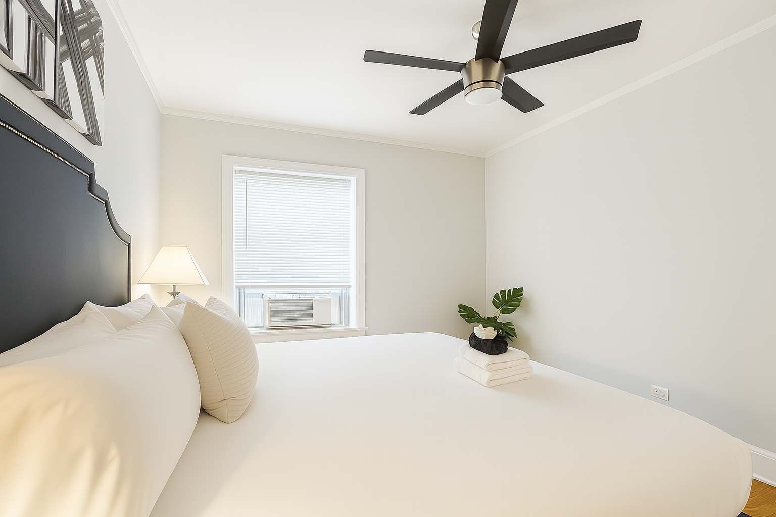 Une chambre à coucher avec un lit, un ventilateur de plafond, une fenêtre avec des stores, une lampe de chevet et une petite plante sur la table de chevet.