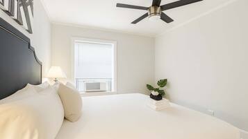 A bedroom with a bed, a ceiling fan, a window with blinds, a bedside lamp, and a small plant on the nightstand.