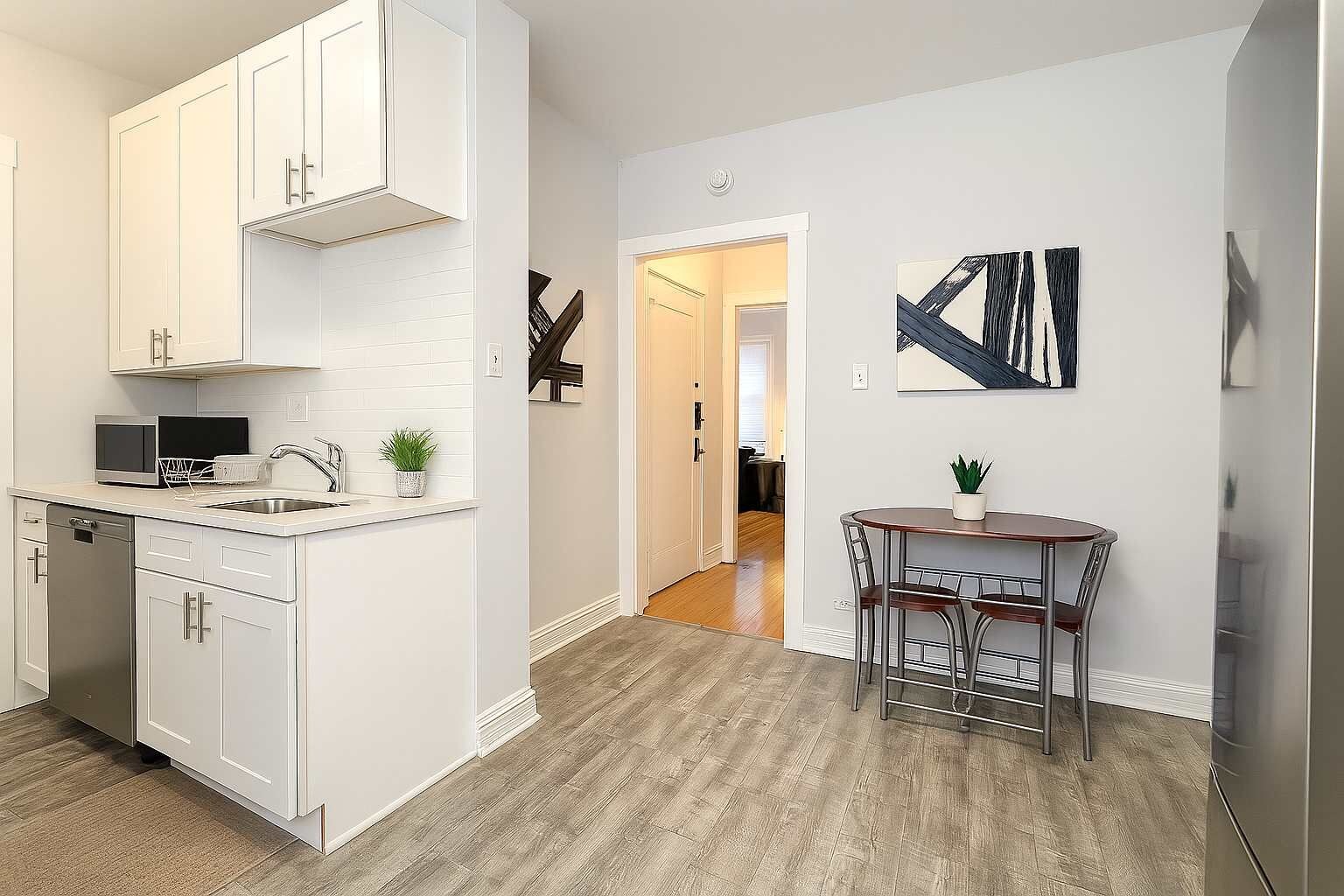Una cocina moderna con muebles blancos, lavavajillas y microondas. Hay un comedor con mesa y sillas, y una escalera que sube a otro nivel.