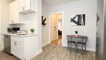 Una cocina moderna con muebles blancos, lavavajillas y microondas. Hay un comedor con mesa y sillas, y una escalera que sube a otro nivel.