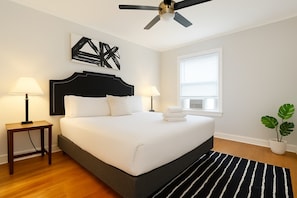 Ein modernes Schlafzimmer mit einem großen Bett, zwei Nachttischlampen, einem Deckenventilator, einem Fenster mit Jalousien und einer Topfpflanze.
