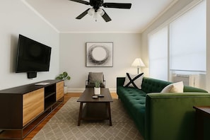 A modern living room with a flat-screen TV, a green sofa, a coffee table, a sideboard, and a ceiling fan.