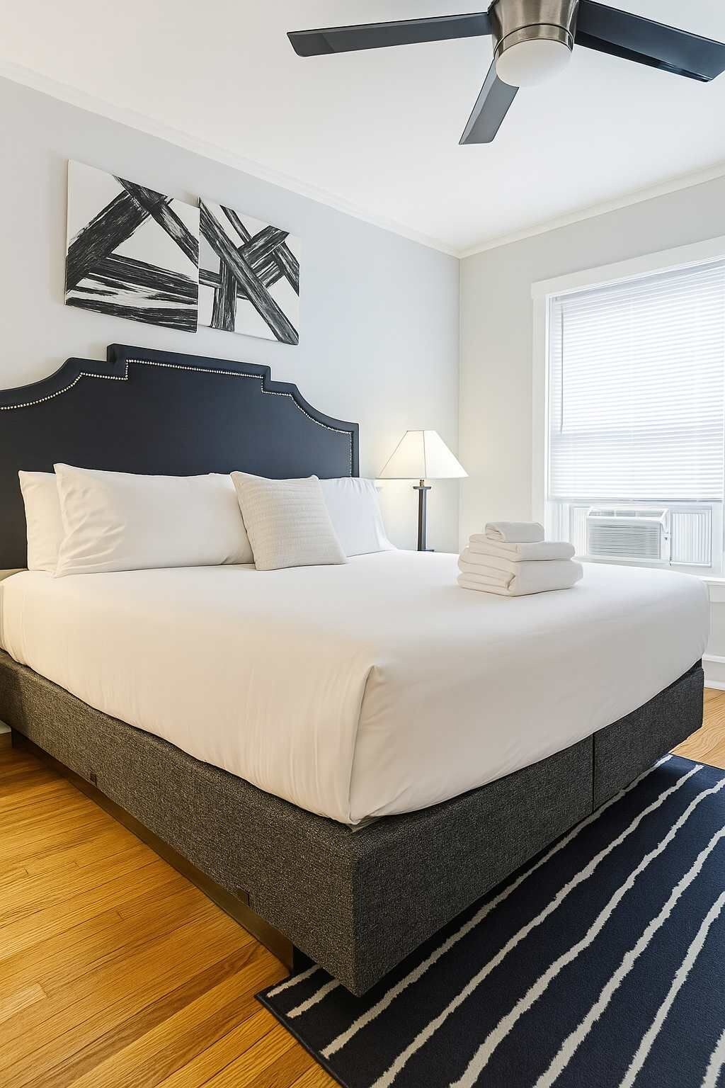 Een keurig opgemaakt bed met witte lakens, een donker hoofdbord en een blauw gestreept vloerkleed.
