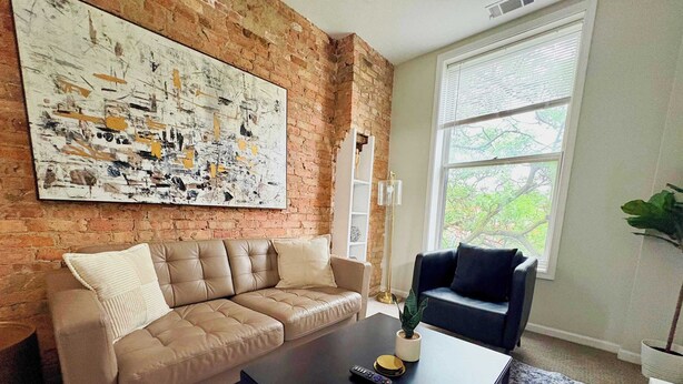 A living room with a brick wall, a large abstract painting, a beige sofa, a dark sofa, a coffee table with a remote control, and a window with a view of trees. - Paradise 1BR Apt in Lincoln Park (Chicago)