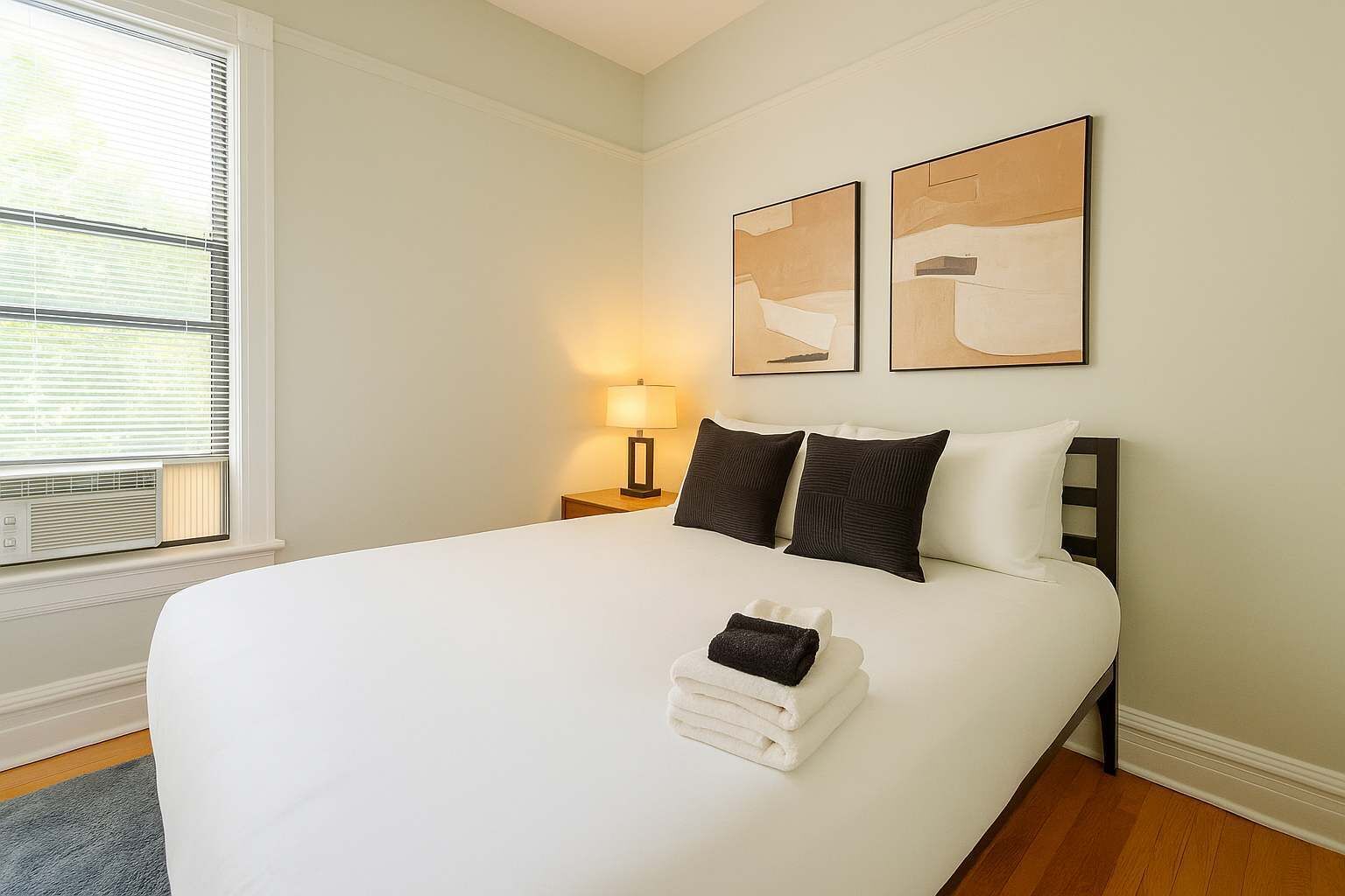A neatly made bed with white linens, a bedside lamp, and two abstract paintings on the wall.